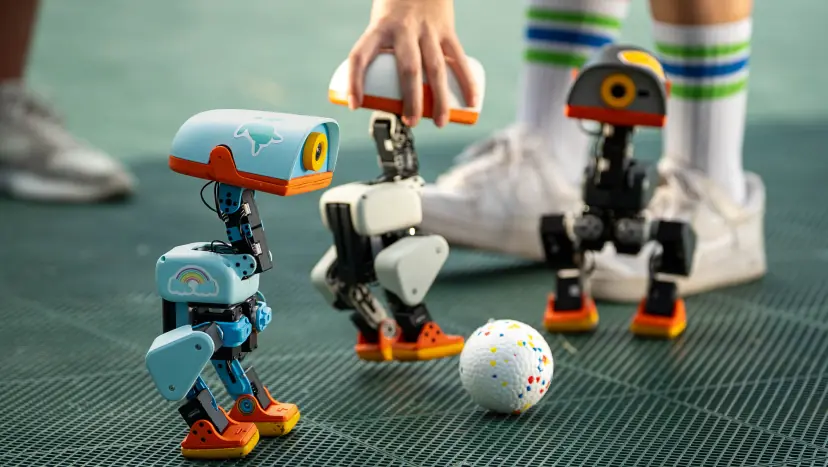 Three Microduck robots beside a ball, with a person steadying one of them.